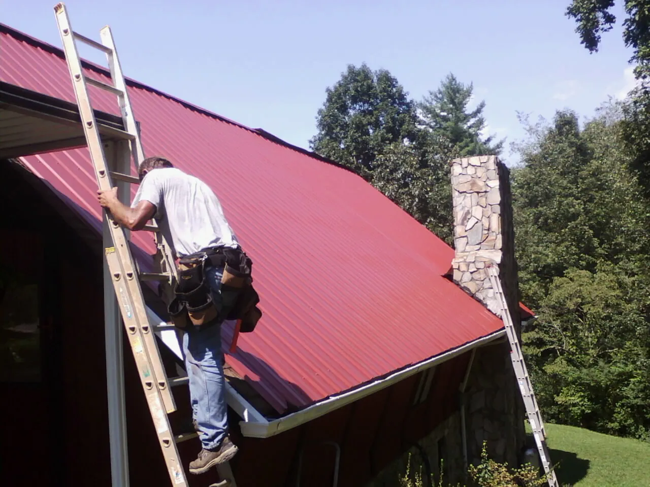 Roofer on a ladder beside a red metal roof and stone chimney — Soffit & Fascia Repair by Roofing Brothers in Moreno Valley